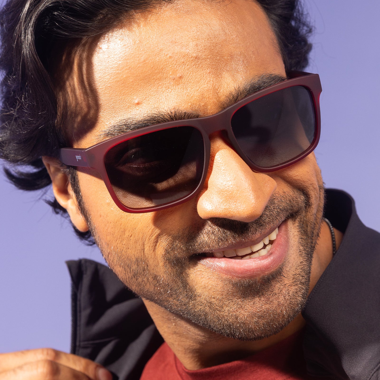 A man with dark wavy hair wearing dark red Grand G frames and black gradient lenses smiles to the side and pops his collar.