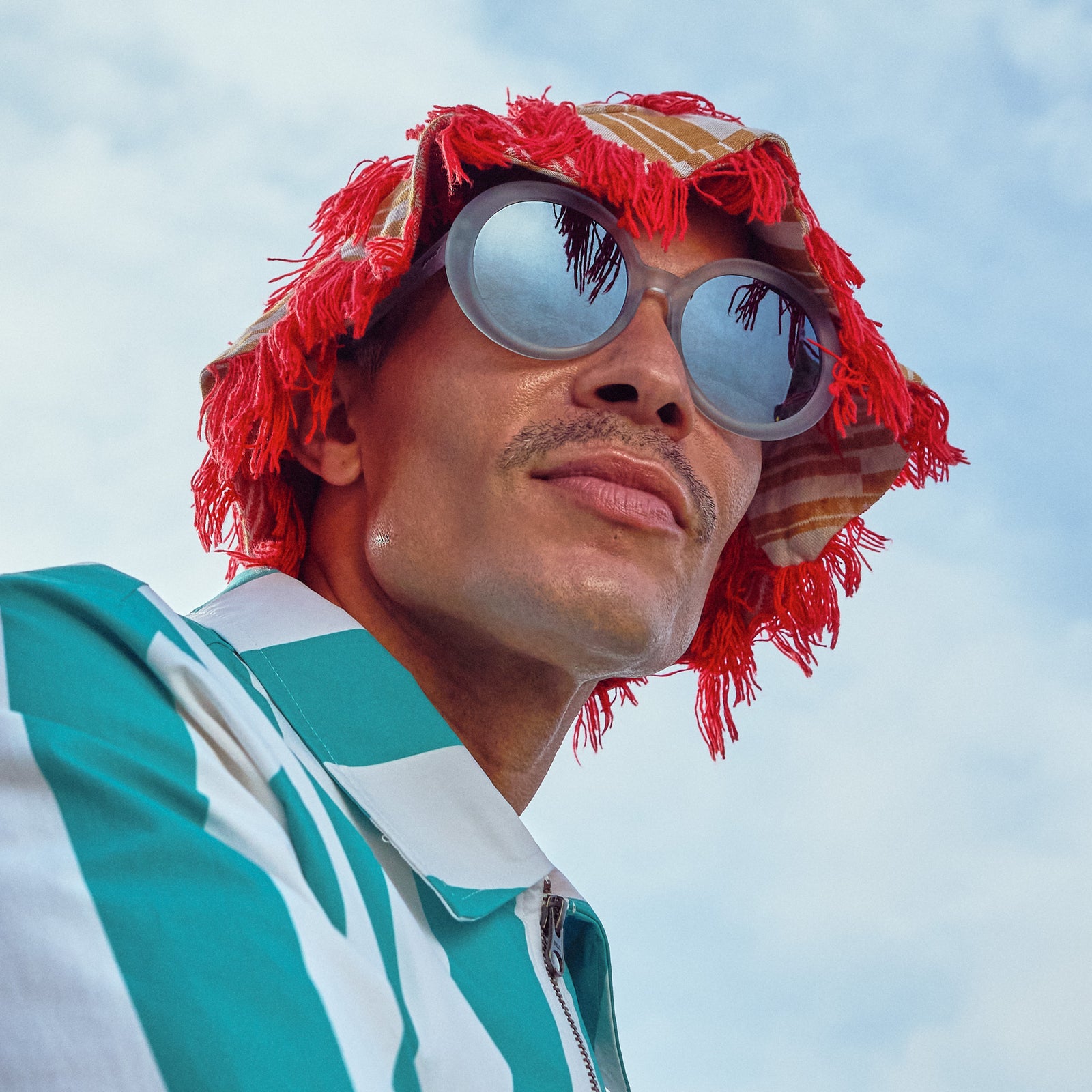 Low-angle view of a man wearing oversized round sunglasses with translucent blue frames and silver reflective gradient lenses, plus a red hat.