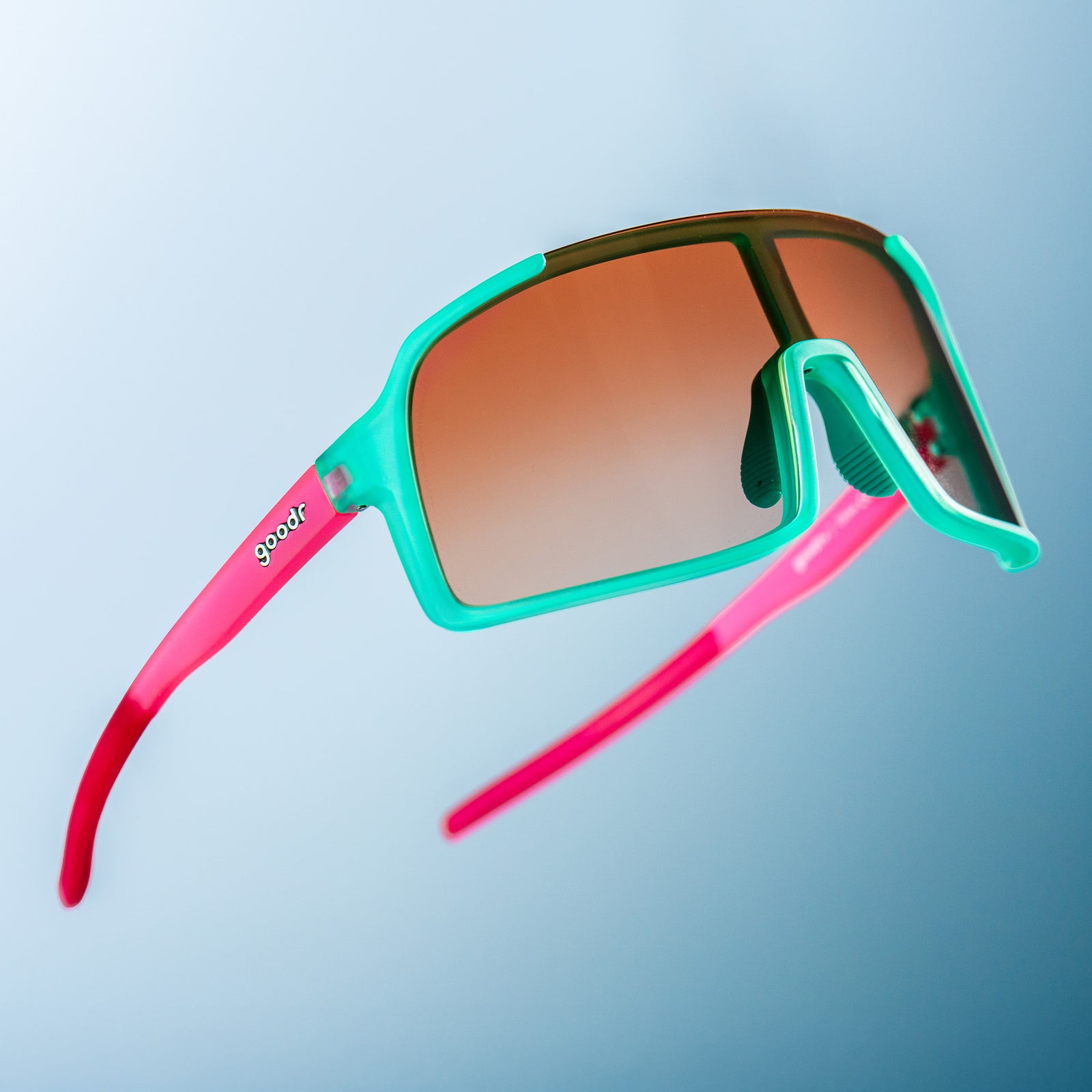 Teal wrap shades with pink arms and amber gradient lenses floating against a blue ombre backdrop.