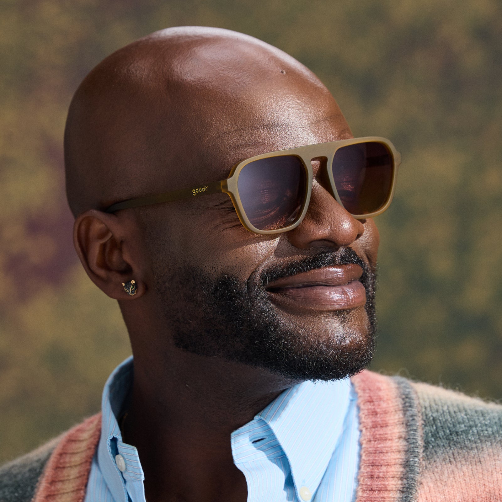 A man smiles in a pair of translucent dusted-ochre Retro G sunglasses against a mottled green backdrop that’s out of focus.