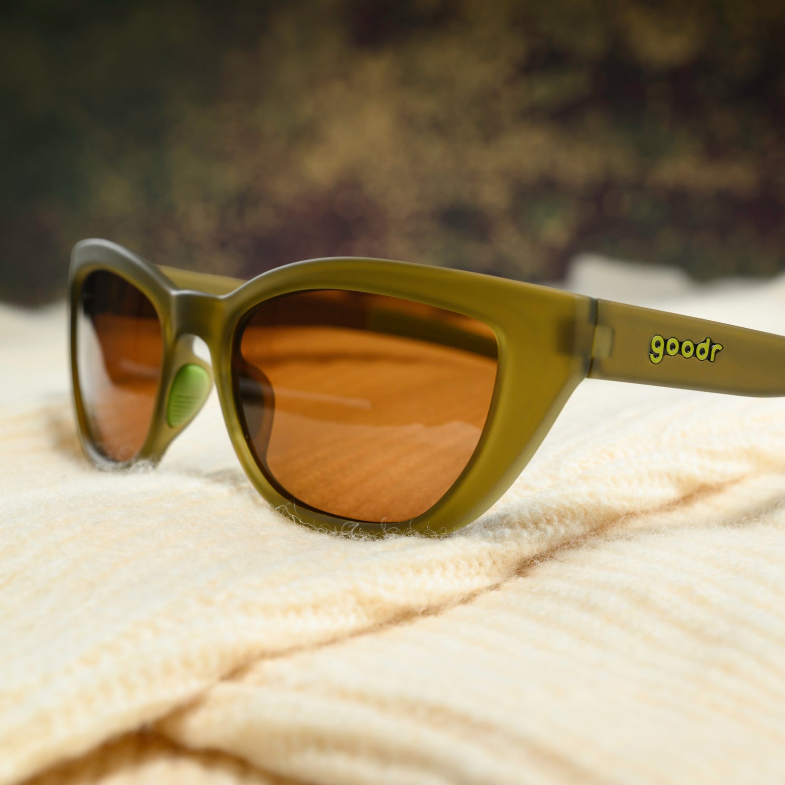 A pair of translucent olive-green cat-eye sunglasses with brown non-reflective lenses sits on a cream-colored sweater.