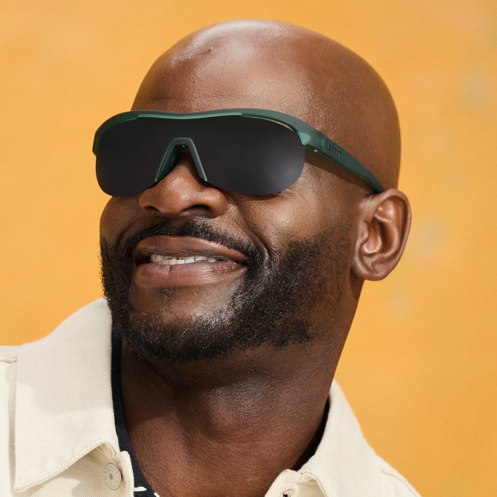 A man wears a pair of translucent dark green Sonic G sunglasses with black non-reflective lenses against a mustard backdrop.