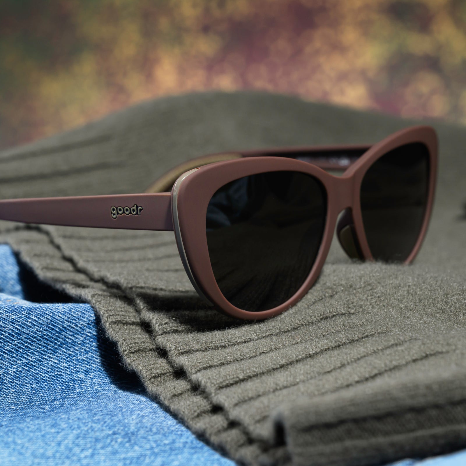 A pair of opaque brown Glam G cat-eye sunglasses with green non-reflective lenses sits atop a green sweater material.