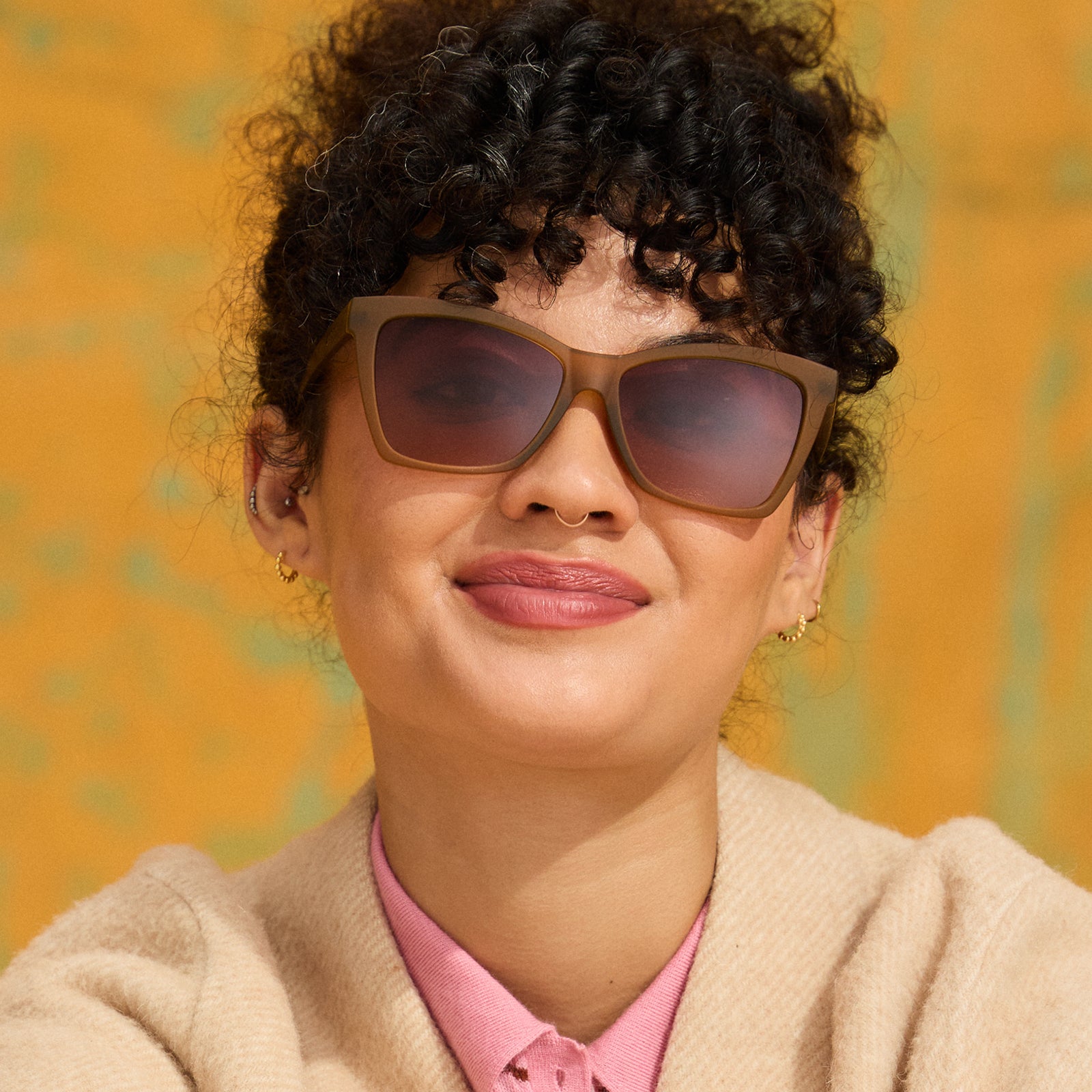 A woman with dark curls smiles warmly in a pair of ochre-yellow angular cat-eye sunnies against a golden ochre background.