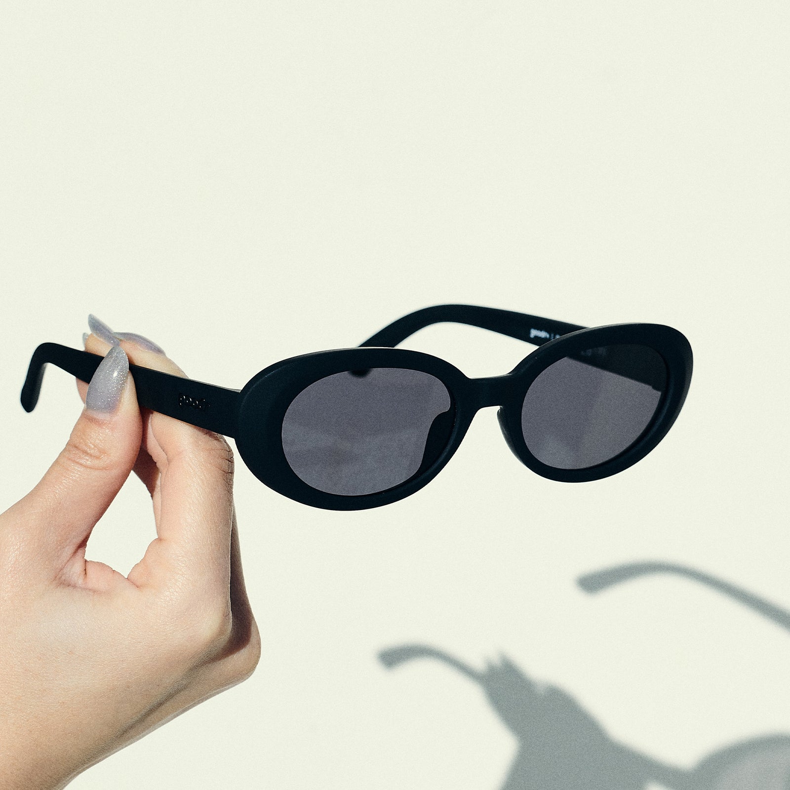 A manicured hand holds a pair of black oval sunglasses with polarized lenses in front of an off-white backdrop.