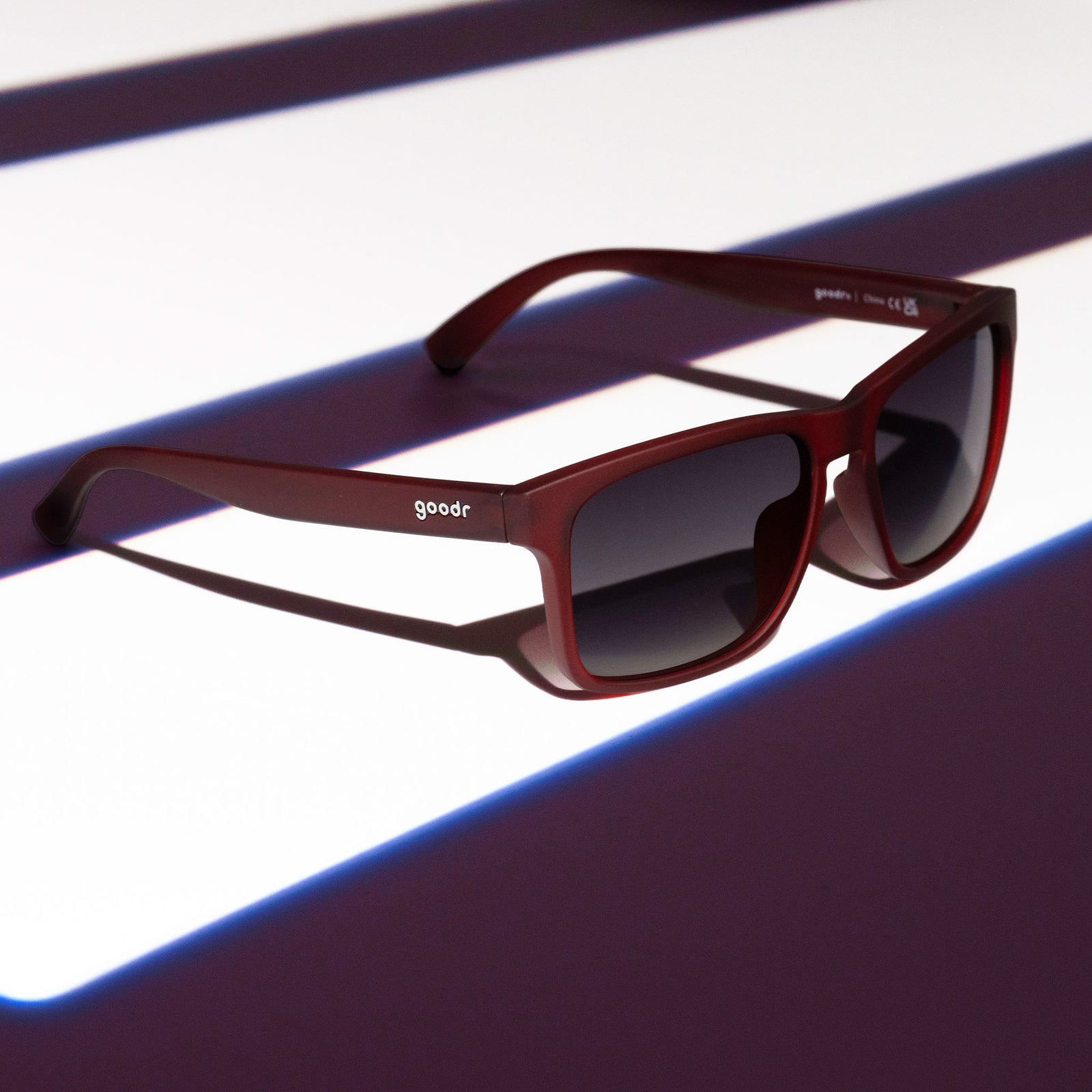 Three-quarter angle view of dark red Grand G frames and black gradient lenses sitting on a white and red striped surface.