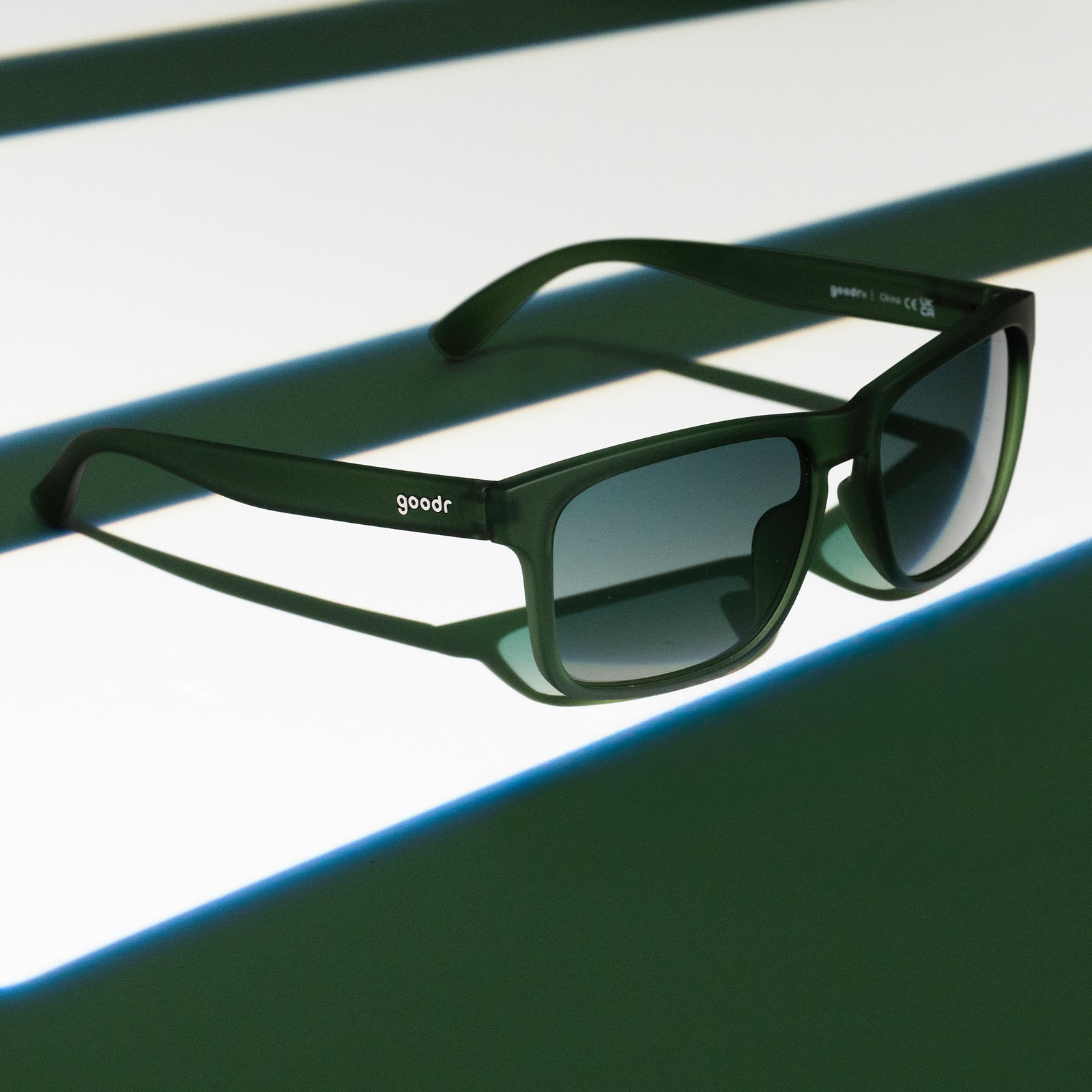 Three-quarter angle view of translucent dark green Grand Gs with green gradient lenses on a dark green striped surface.