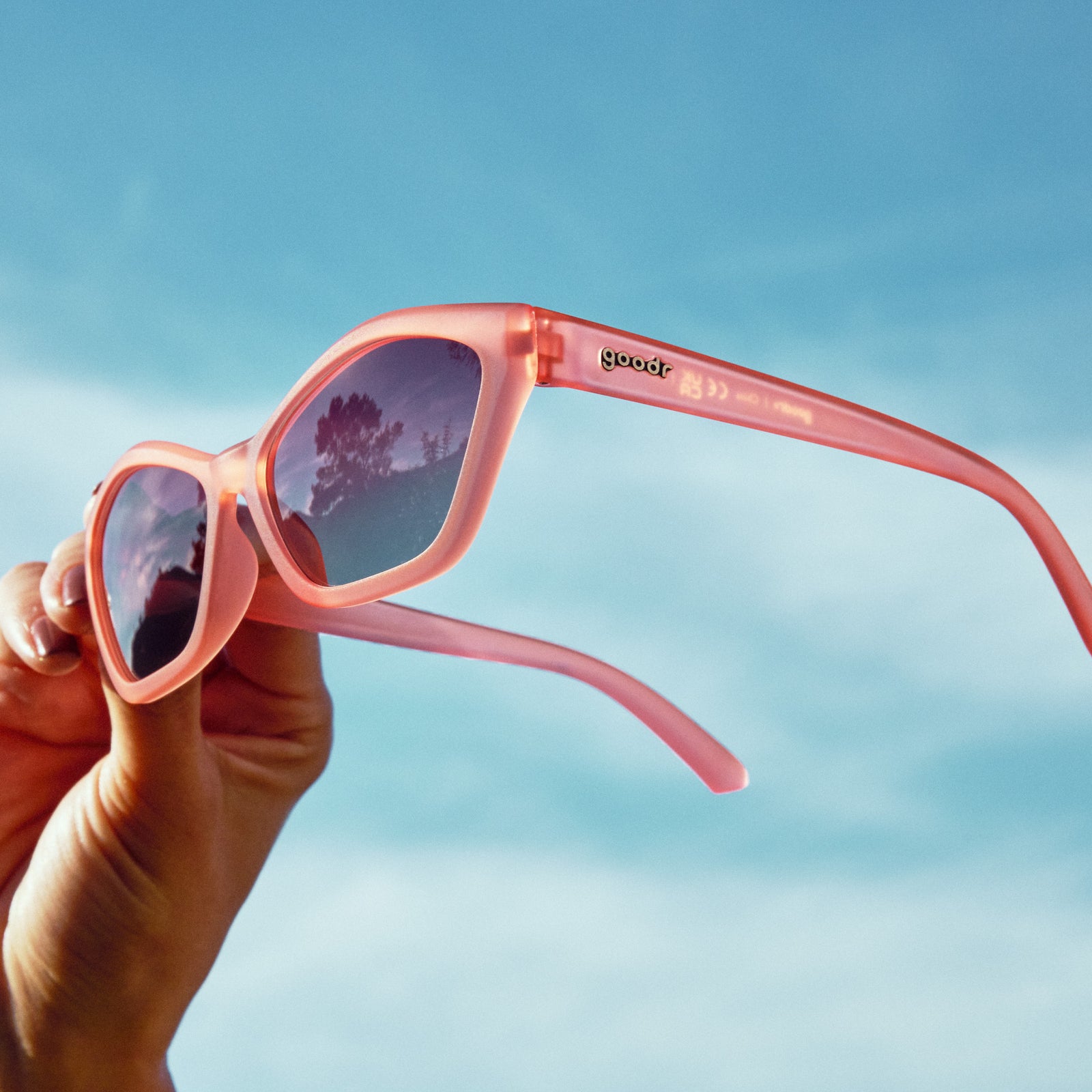 A manicured hand holds a pair of geometric sunglasses with translucent coral pink frames and gradient lenses against the sky at a three-quarter angle.