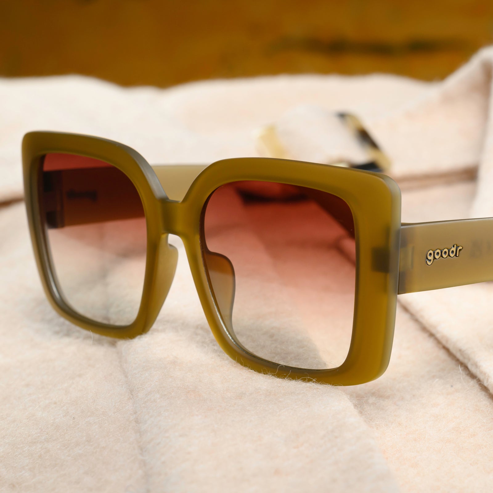 Translucent dusted-ochre, oversized square Amp G sunglasses sitting on a woolly cream-colored jacket sleeve. 