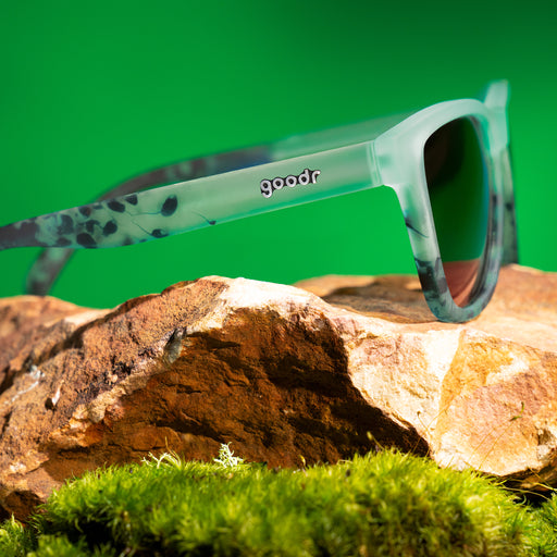 Three-quarter angle view of translucent green sunglasses with dark green hand-painted swirls and gradient lenses on a rock.