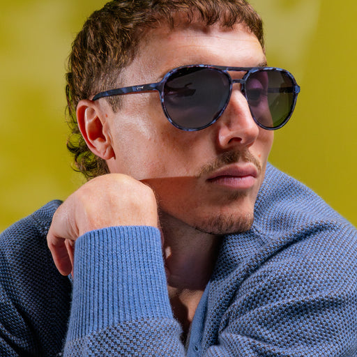 A man posing while wearing translucent blue tortoiseshell aviator frames featuring black gradient lenses.