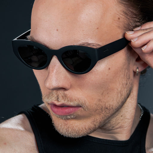 Close-up of a man’s left side profile wearing translucent black, retro-inspired angled cat-eye sunglasses with non-reflective lenses.