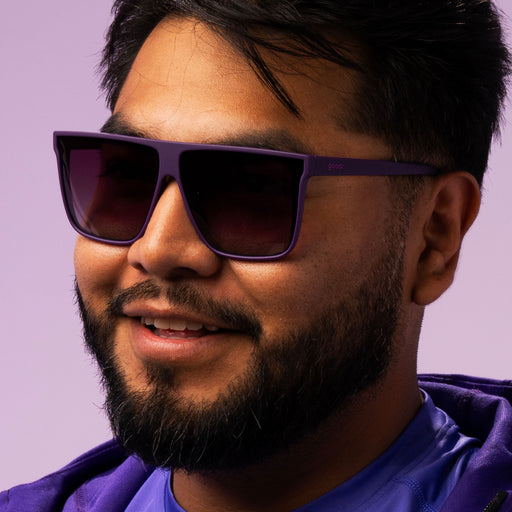 A closeup of a smiling man in a purple shirt vibing in his oversized dark purple flat-top sunglasses.