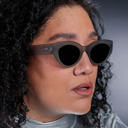 Close-up of a woman's left side profile, wearing translucent dark gray, retro-inspired angled cat-eye sunglasses with black non-reflective lenses.