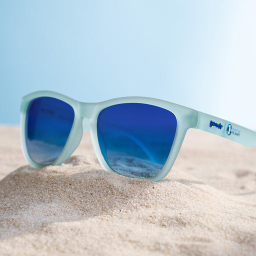 Three-quarter angle view of light blue sunglasses perched atop a small mound of sand against a sky-blue backdrop.