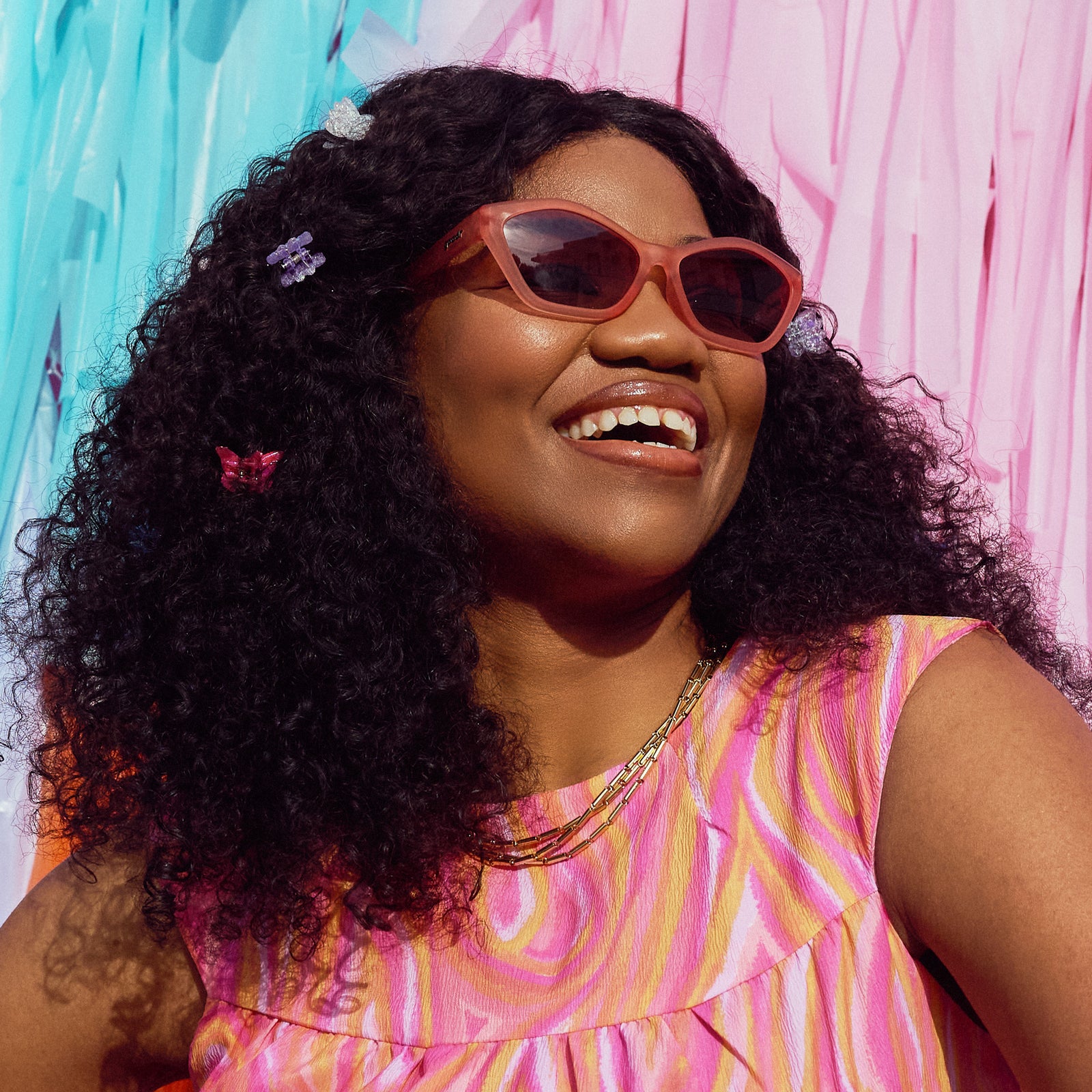 A smiling woman wearing geometric sunglasses with translucent coral pink frames and gradient lenses in front of a pastel backdrop.