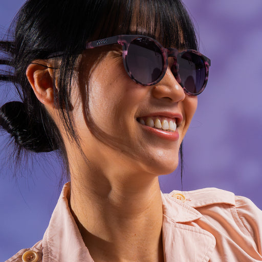 A woman smiling while wearing round translucent pink tortoiseshell frames with black gradient lenses.