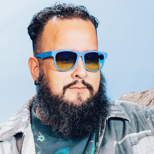 Front view of a man posing in front of a mountain backdrop wearing large blue glasses with blue-yellow ocean gradient lenses.