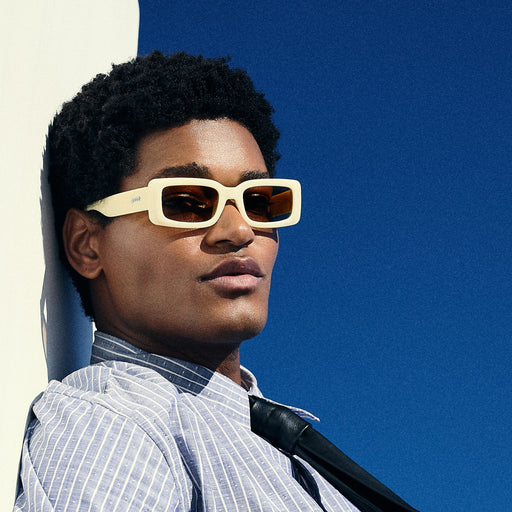 A man leans against a wall wearing a pair of yellow rectangular sunglasses with brown-tinted polarized lenses.