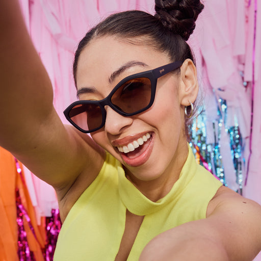 Woman smiles and poses wearing black geometric cat-eye sunglasses with amber gradient lenses against a colorful background.