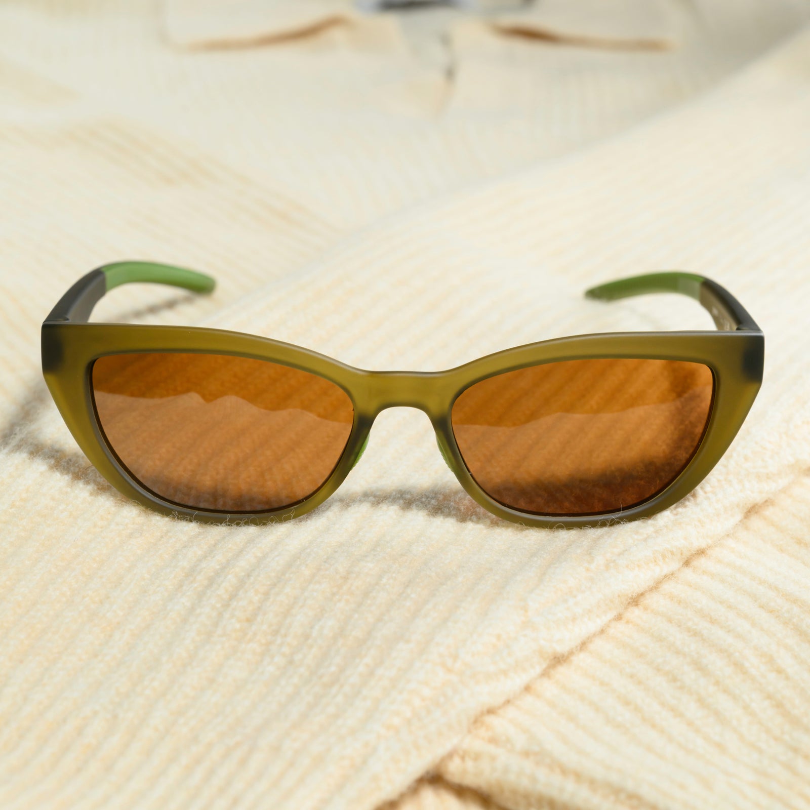 A pair of translucent olive-green cat-eye sunglasses with brown non-reflective lenses sits on a cream-colored sweater.