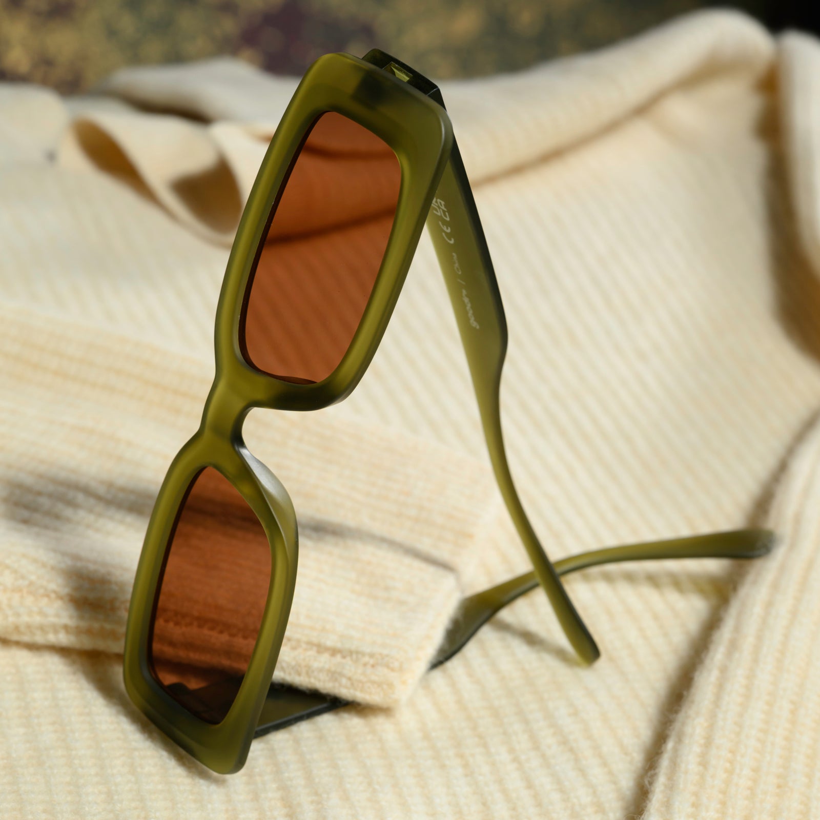 A pair of translucent, rectangular olive-green sunglasses with brown non-reflective lenses sits on a cream-colored sweater.