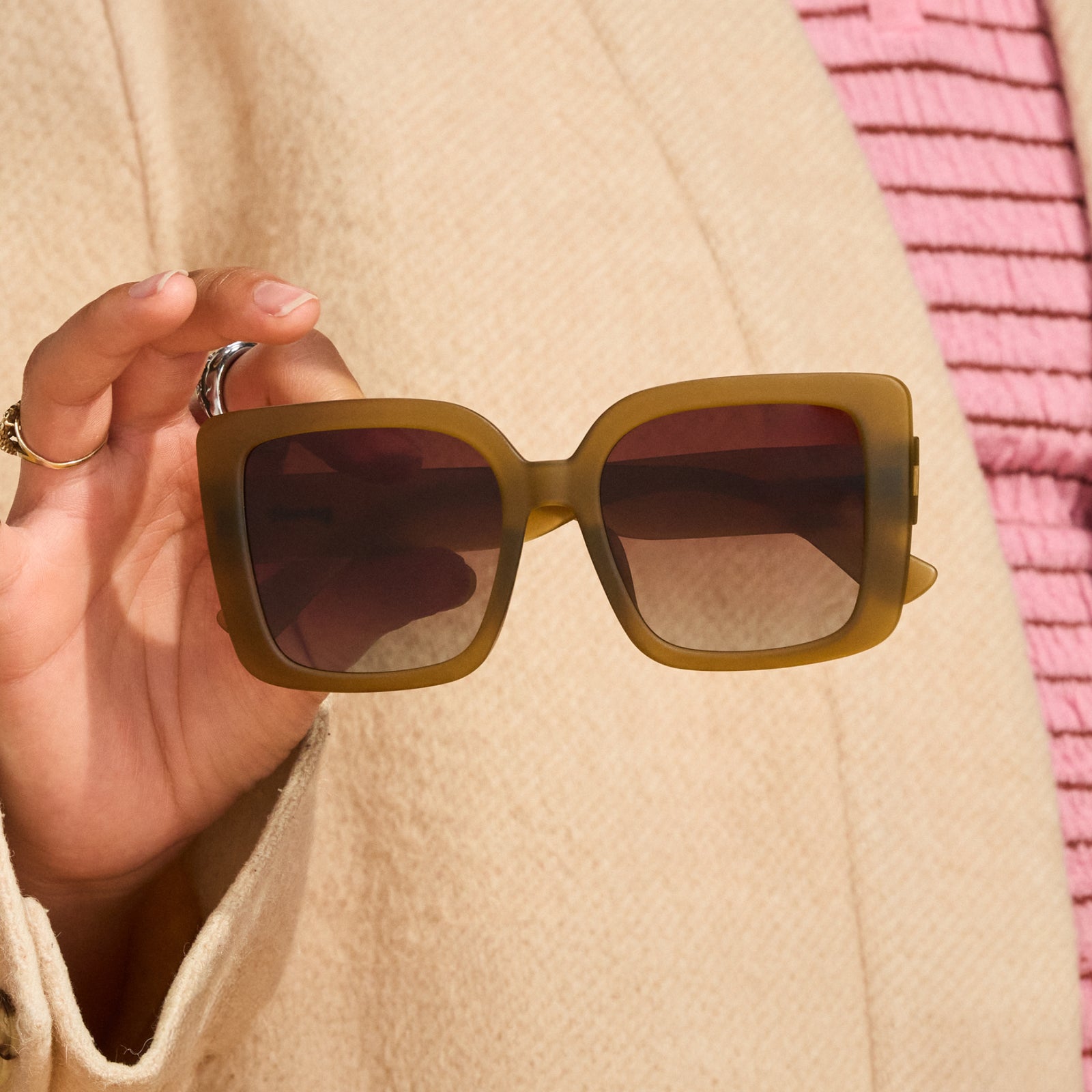 Close-up of a woman’s hand holding a pair of dusted-ochre, square Amp G sunglasses in front of her torso.