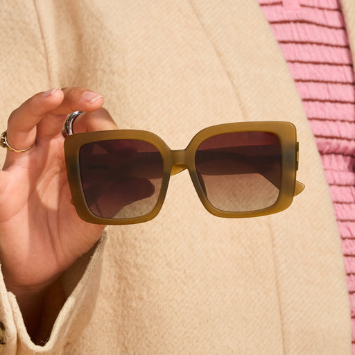 Close-up of a woman’s hand holding a pair of dusted-ochre, square Amp G sunglasses in front of her torso.