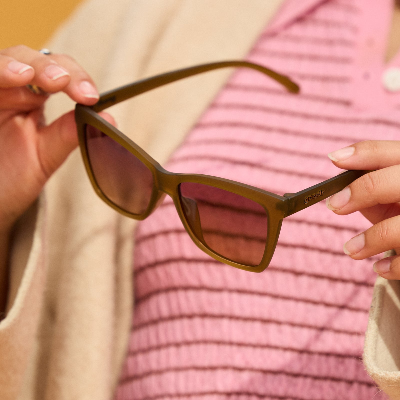 Close-up of a woman’s hands holding a pair of translucent ochre sunglasses in front of her pink and cream outfit.