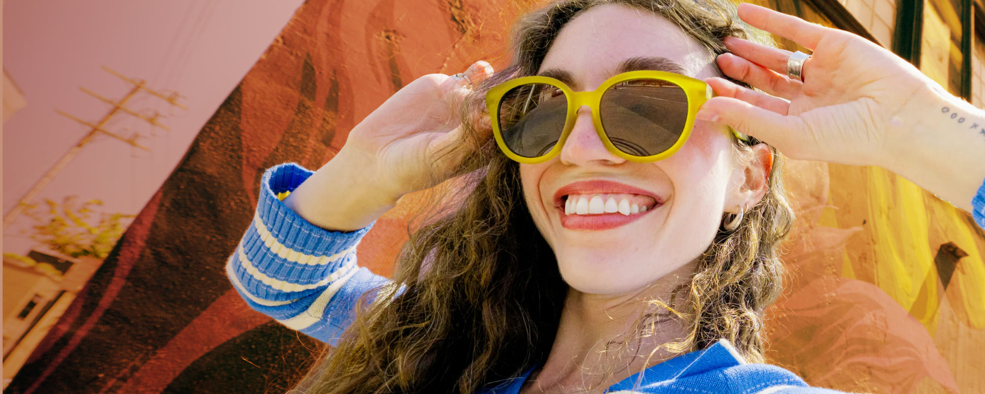 A smiling woman wearing bright yellow square sunglasses with dark lenses, holding them in place with both hands against a colorful outdoor background.
