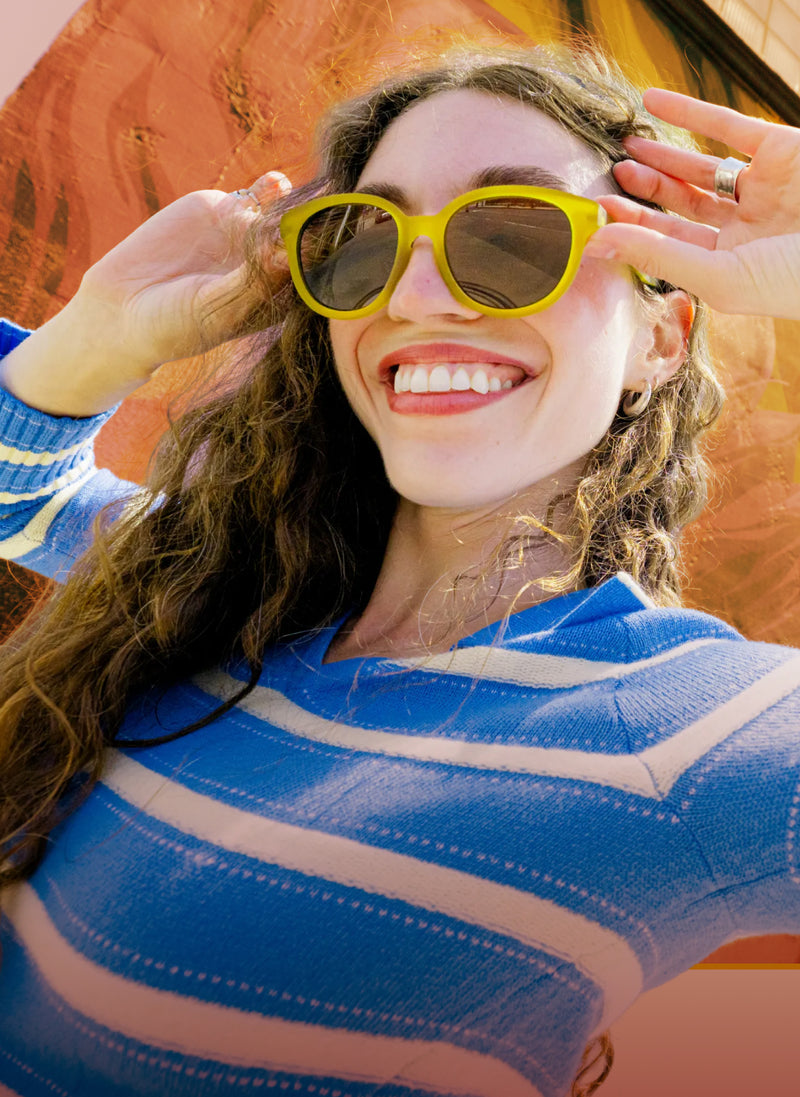 A smiling woman with long brown hair wearing bright yellow cat-eye sunglasses and a blue and white striped knit sweater, posing in front of a warm orange-brown textured background.