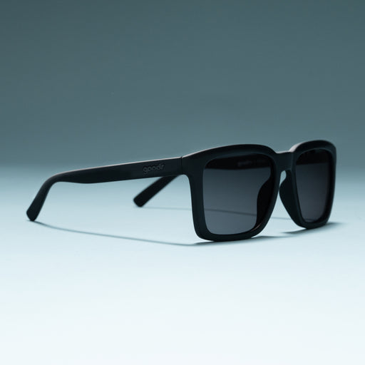 Three-quarter angle view of black shades w/ black nonreflective lenses+ a classic square silhouette on a flat surface.