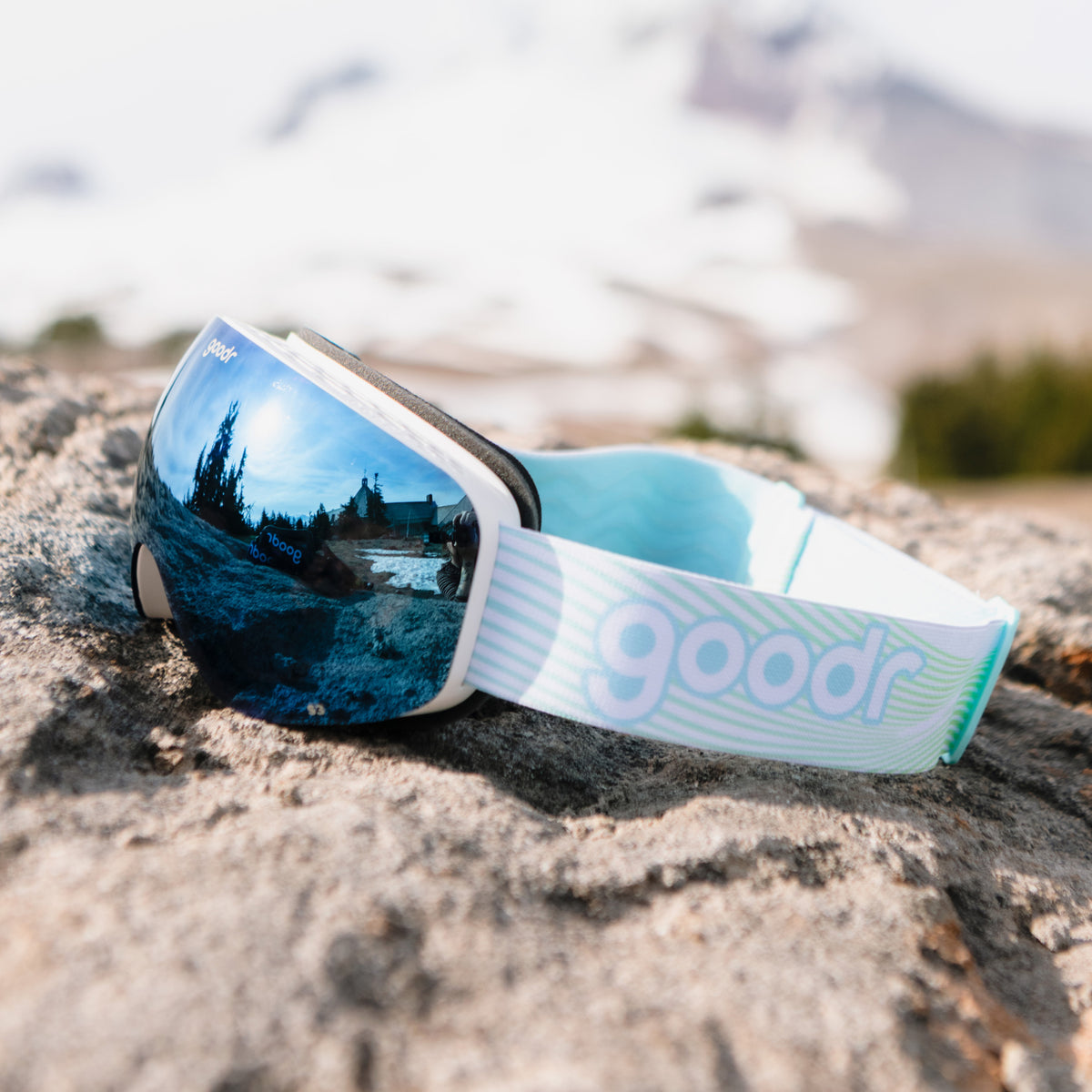 Blue mirrored snow goggles with a white frame and white goodr strap resting on a rock.