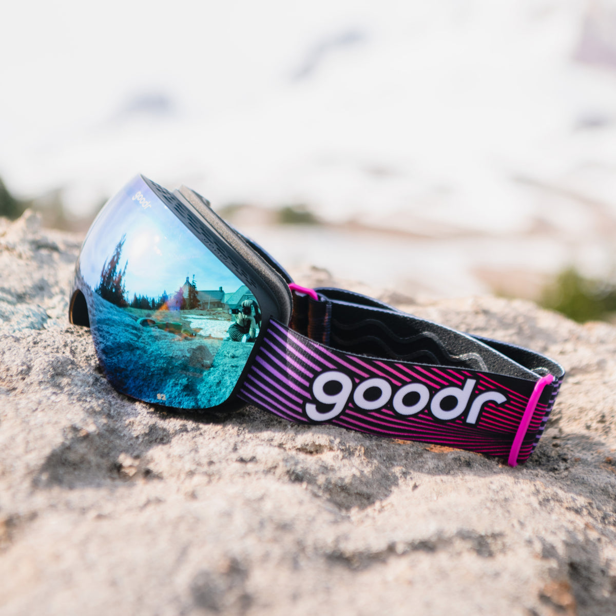 Bronze blue mirrored snow goggles with a black frame and pink and purple goodr strap resting on a rock.
