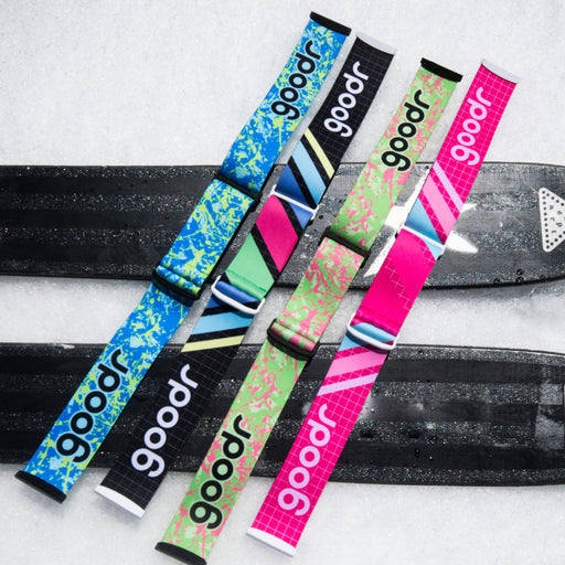 Four colorful goodr replacement goggle straps arranged across black skis in snow.