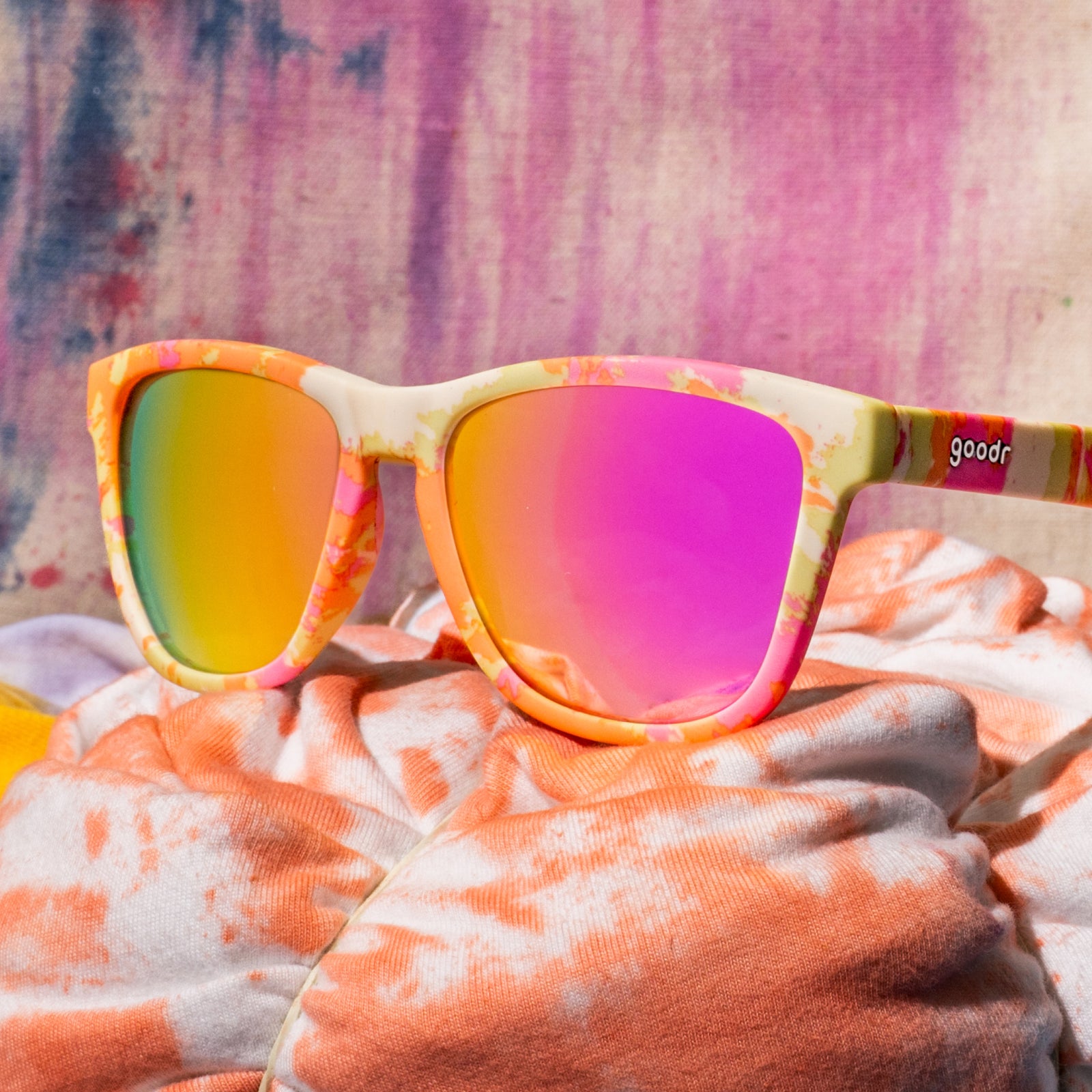 Yellow, pink, and orange tie-dye shades with pink reflective lenses sitting on white and orange tie-dyed fabric.