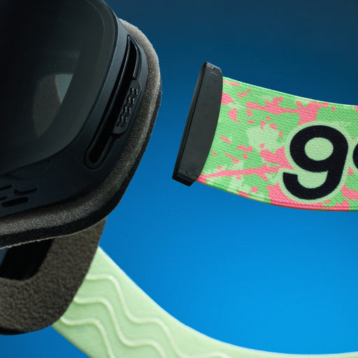 Close-up of green and pink splatter goodr goggle strap attached to black ski goggles.