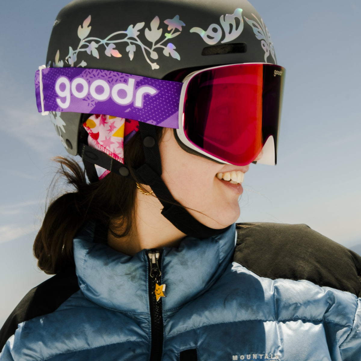 Skier wearing purple red mirrored snow goggles with a purple goodr strap and black helmet.