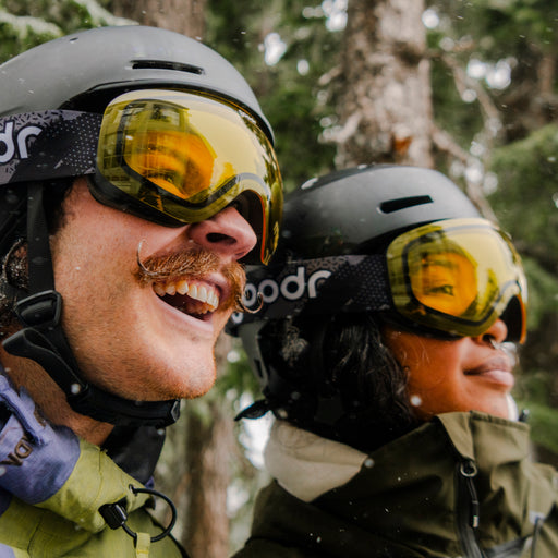 Two skiers wearing yellow snow goggles with patterned goodr straps and black helmets.