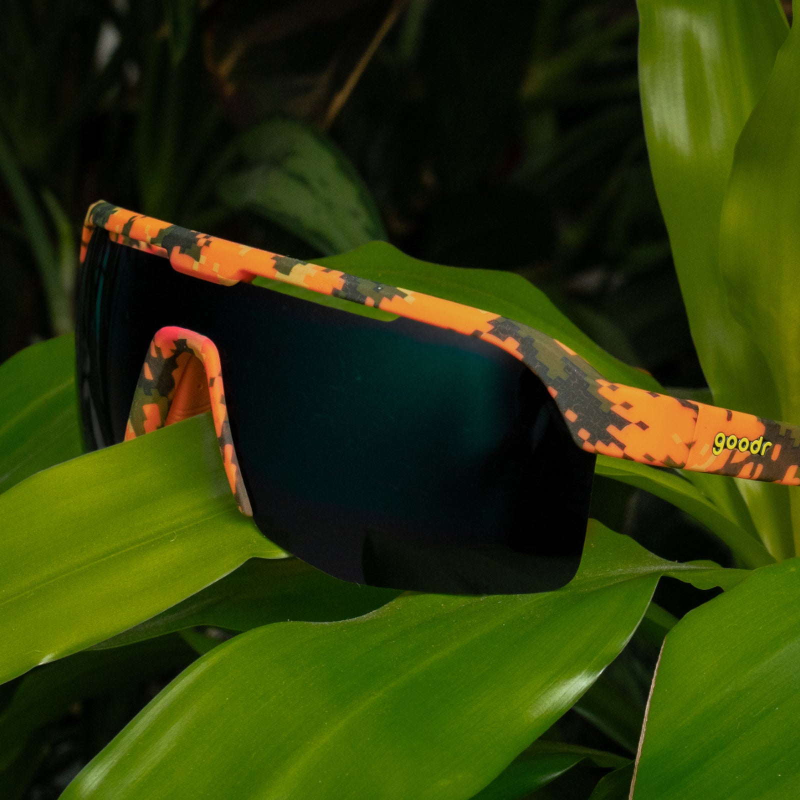 Orange camouflage sport sunglasses with dark mirrored lenses resting on bright green tropical leaves. The brand name 'goodr' is printed in yellow on the temple arm.