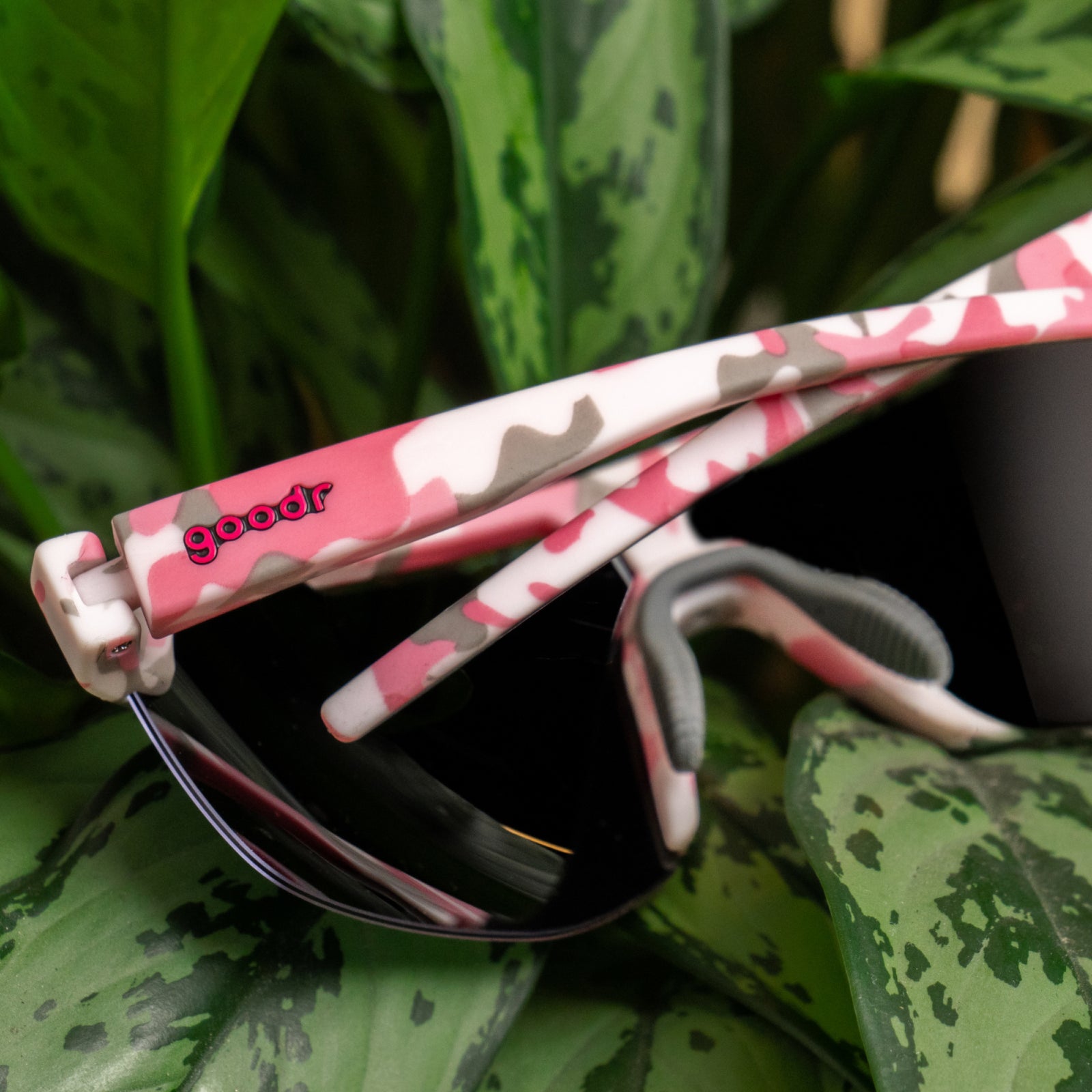Pink, white, and gray camouflage sunglasses with a visible 'goodr' logo on the temple, resting on green patterned leaves.