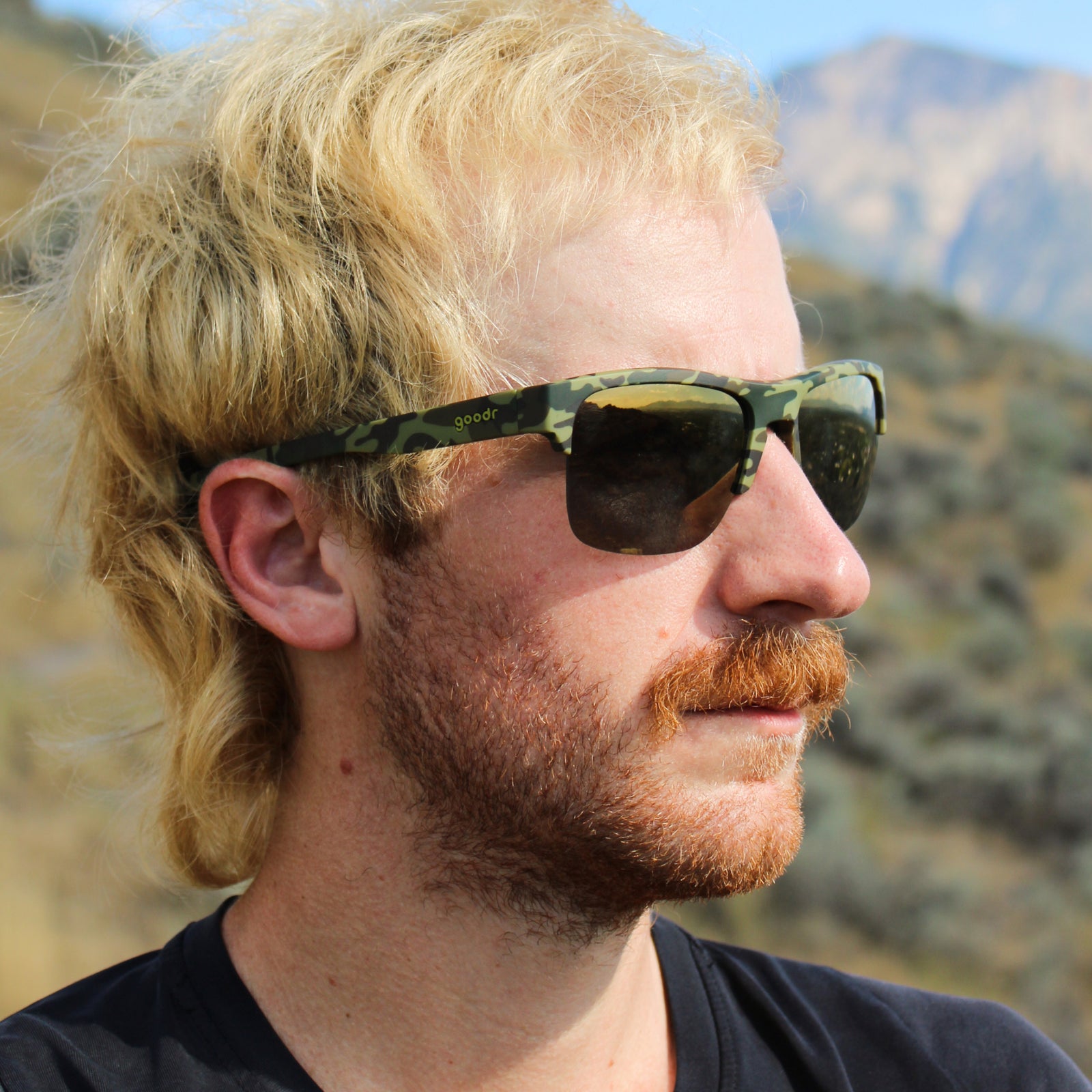 A close-up side profile of a man with blonde hair and a reddish beard wearing green camouflage sunglasses, set against a blurred outdoor mountain background.