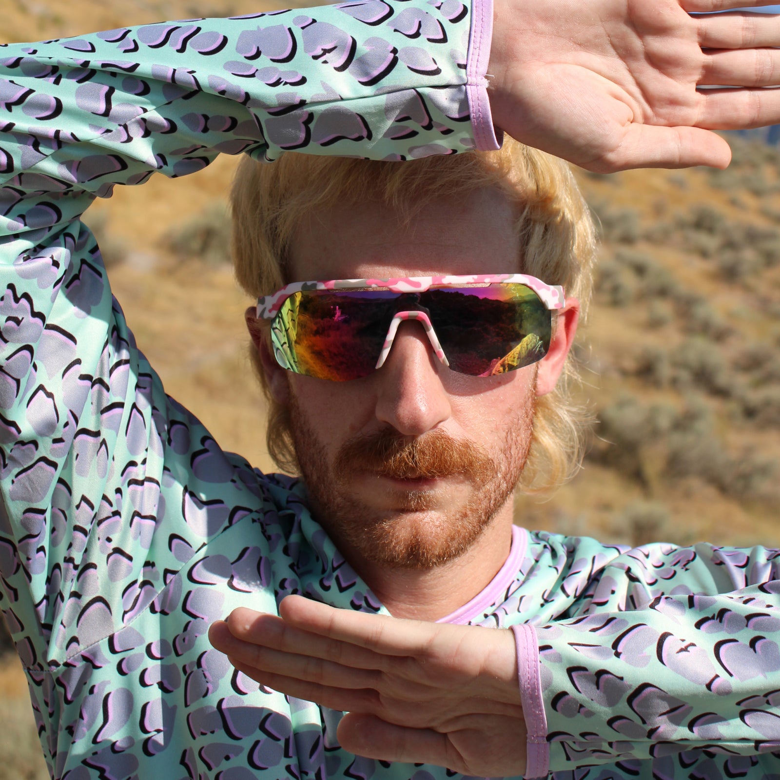 A close-up lifestyle shot of a man outdoors wearing a mint green and purple patterned long-sleeve athletic shirt and mirrored sunglasses.