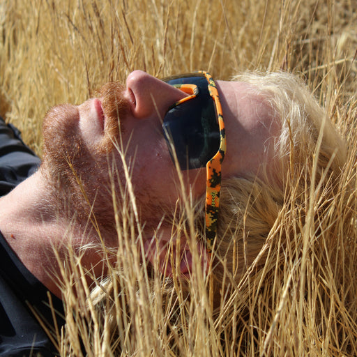 A close-up lifestyle shot of a man lying in a field of dry golden grass, wearing black sunglasses with a vibrant orange and black patterned temple arm.